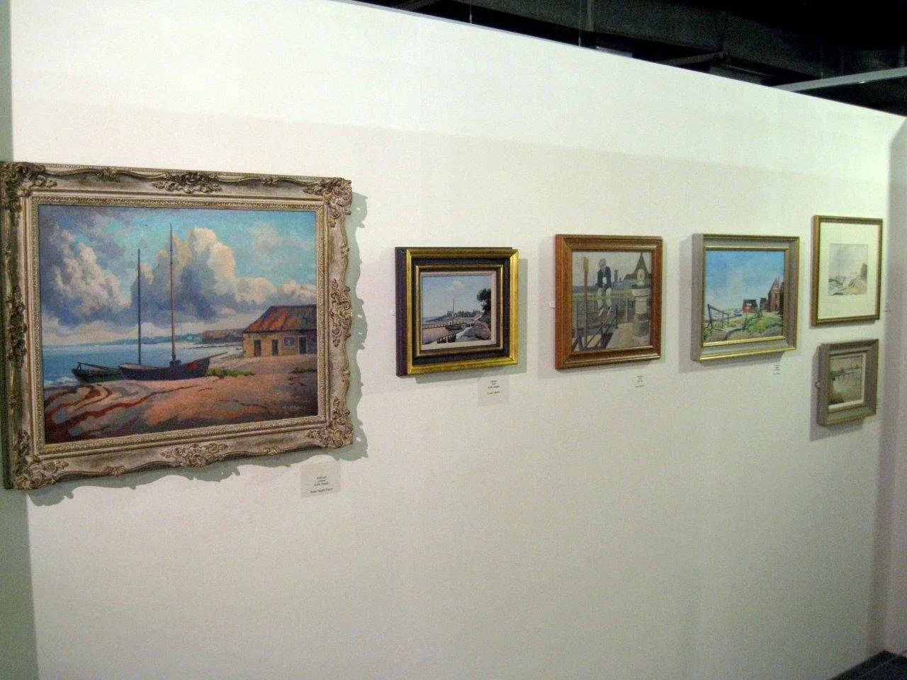 A series of framed landscape paintings displayed on a white gallery wall, with the largest painting on the left featuring boats, a house, and a cloudy sky.