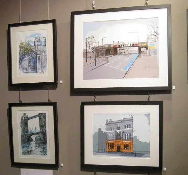 Four framed watercolor paintings hung on a gallery wall, two on top and two below, depicting urban scenes including a bridge, a street under a bridge, a historic building, and a church.