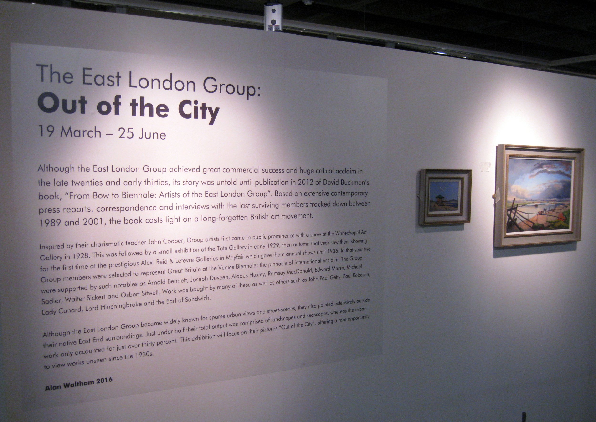 Exhibition panel titled 'The East London Group: Out of the City,' with display dates from March 19 to June 25. The panel contains a paragraph of text explaining the achievement and history of the East London Group, a group of artists who gained prominence in the early 20th century. It also features two small framed paintings of landscapes on the right side of the panel.