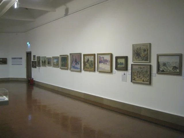 Gallery wall with multiple landscape paintings in a museum or gallery setting.