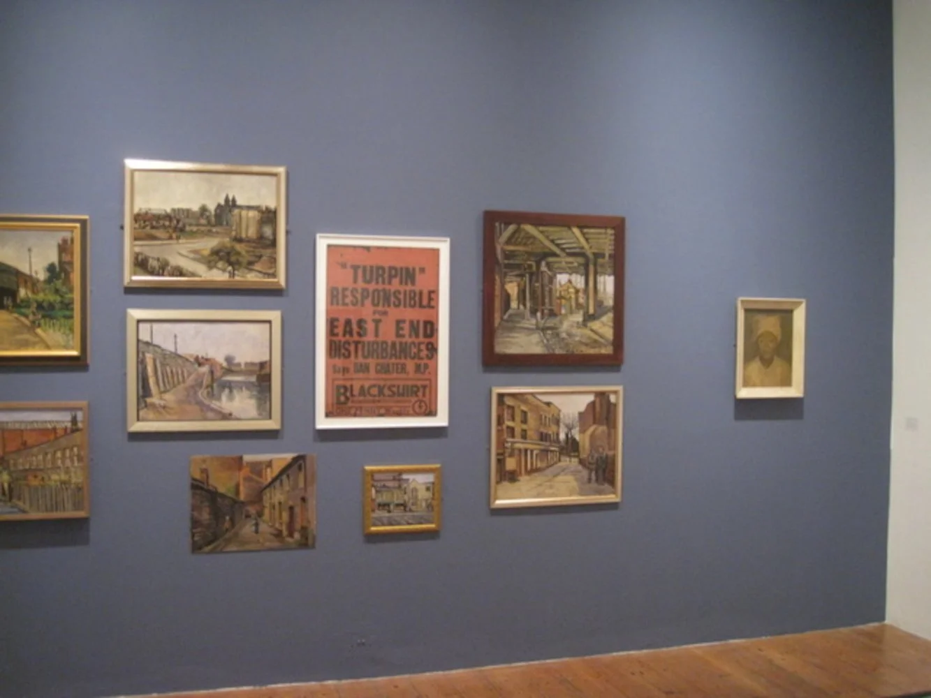 Gallery wall with various paintings and a sign, blue wall background, wooden floor.