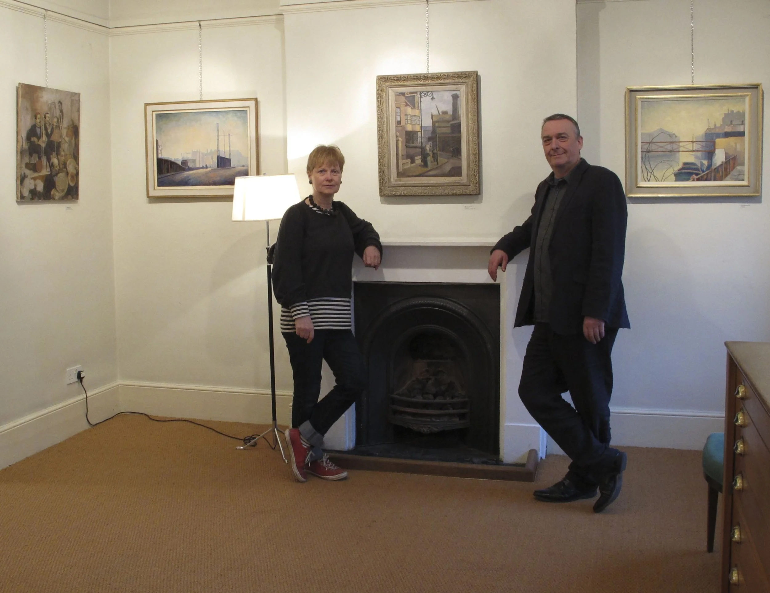 A woman and a man standing beside a fireplace in a gallery or art exhibition space, with four framed paintings on the wall above the fireplace and a floor lamp nearby.