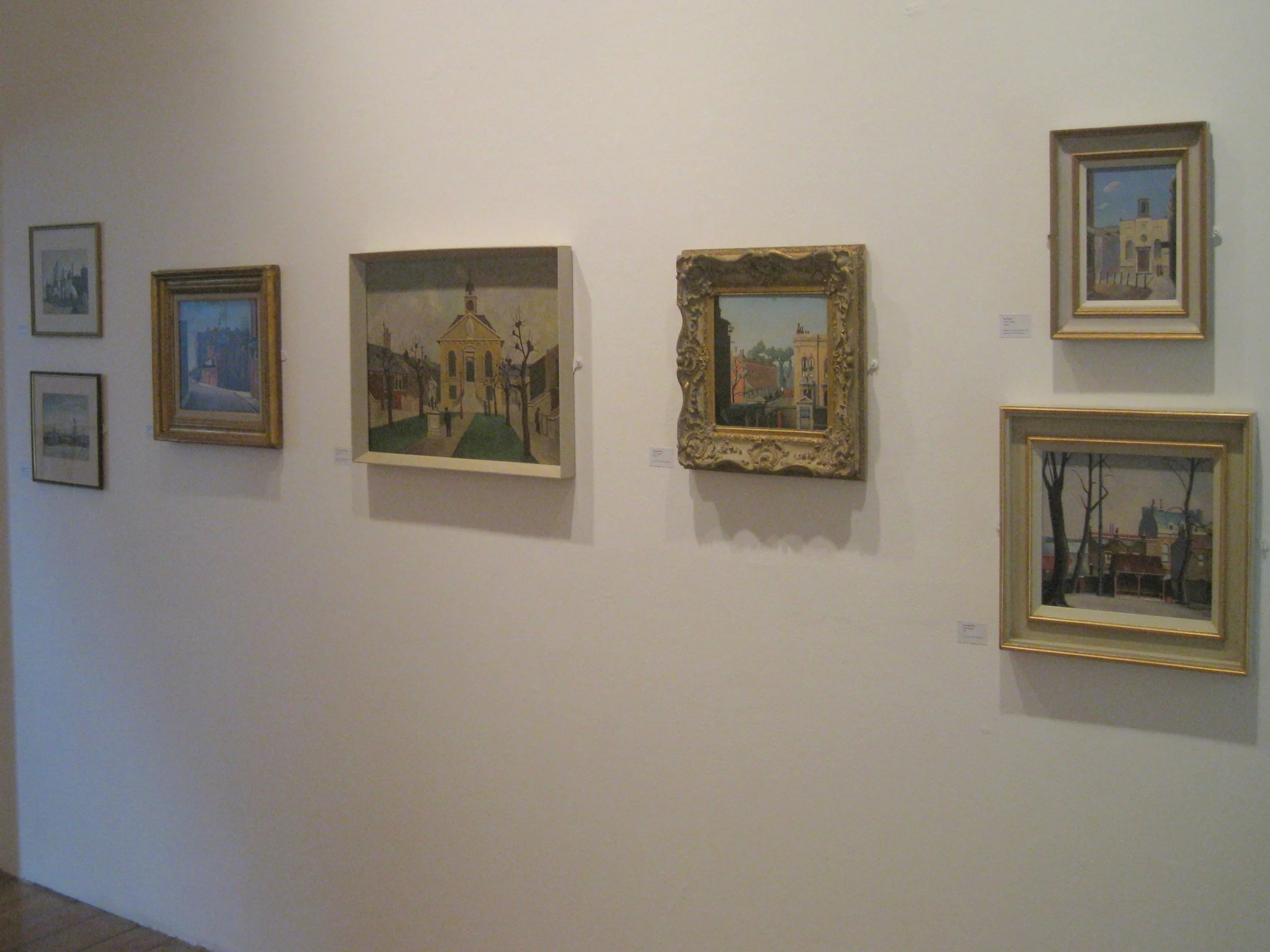 Gallery wall with eight framed paintings depicting cityscapes, buildings, and landscapes in various styles and sizes.