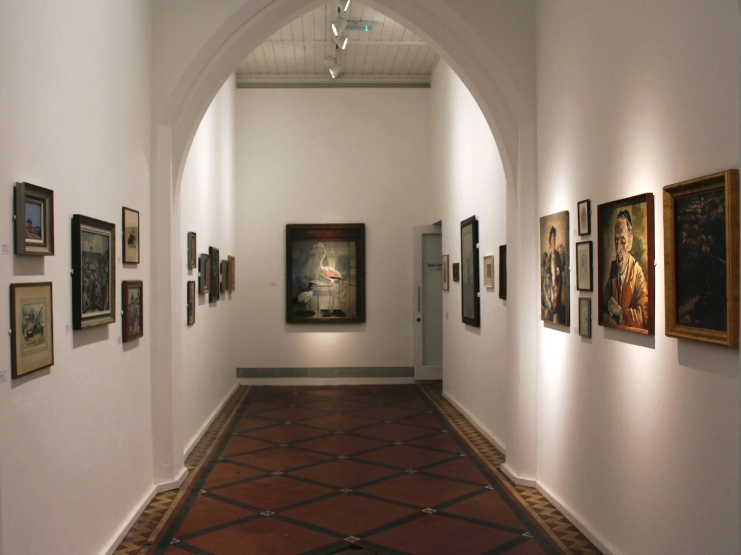 Art gallery with framed paintings on white walls and a geometric-patterned wooden floor.