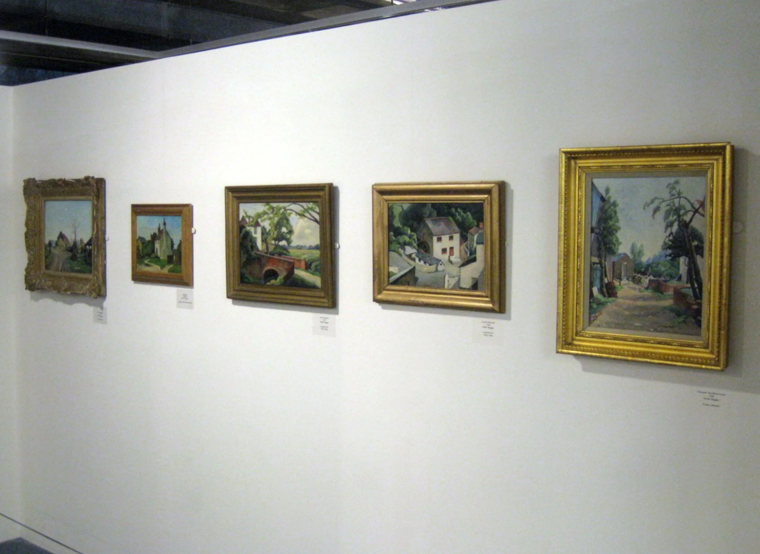 A gallery wall displaying six landscape paintings in various frames, featuring rural scenes with trees, houses, bridges, and paths.