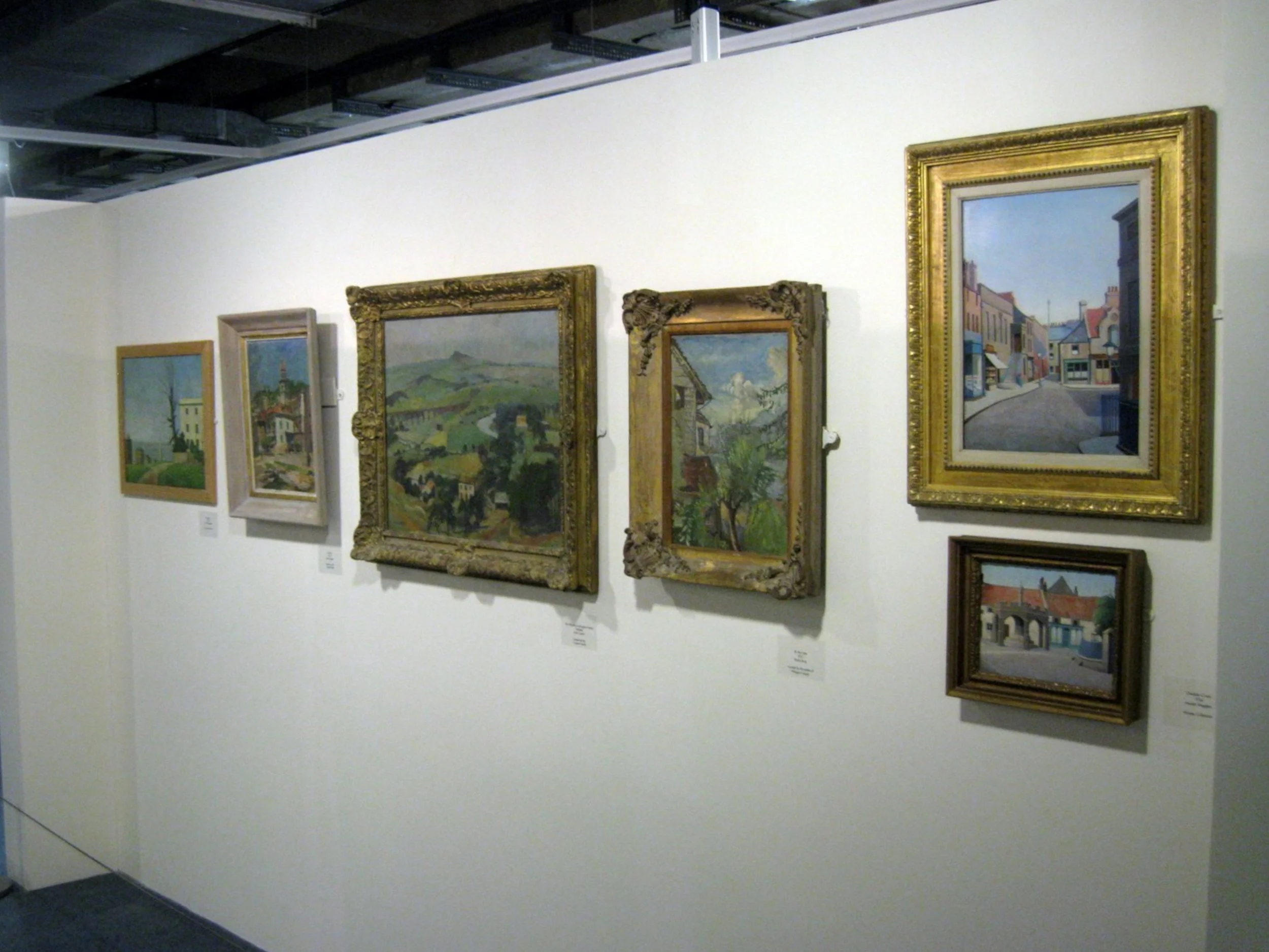 A display of seven framed landscape paintings on a white gallery wall, with some frames ornate and others simple, depicting rural and urban scenes.