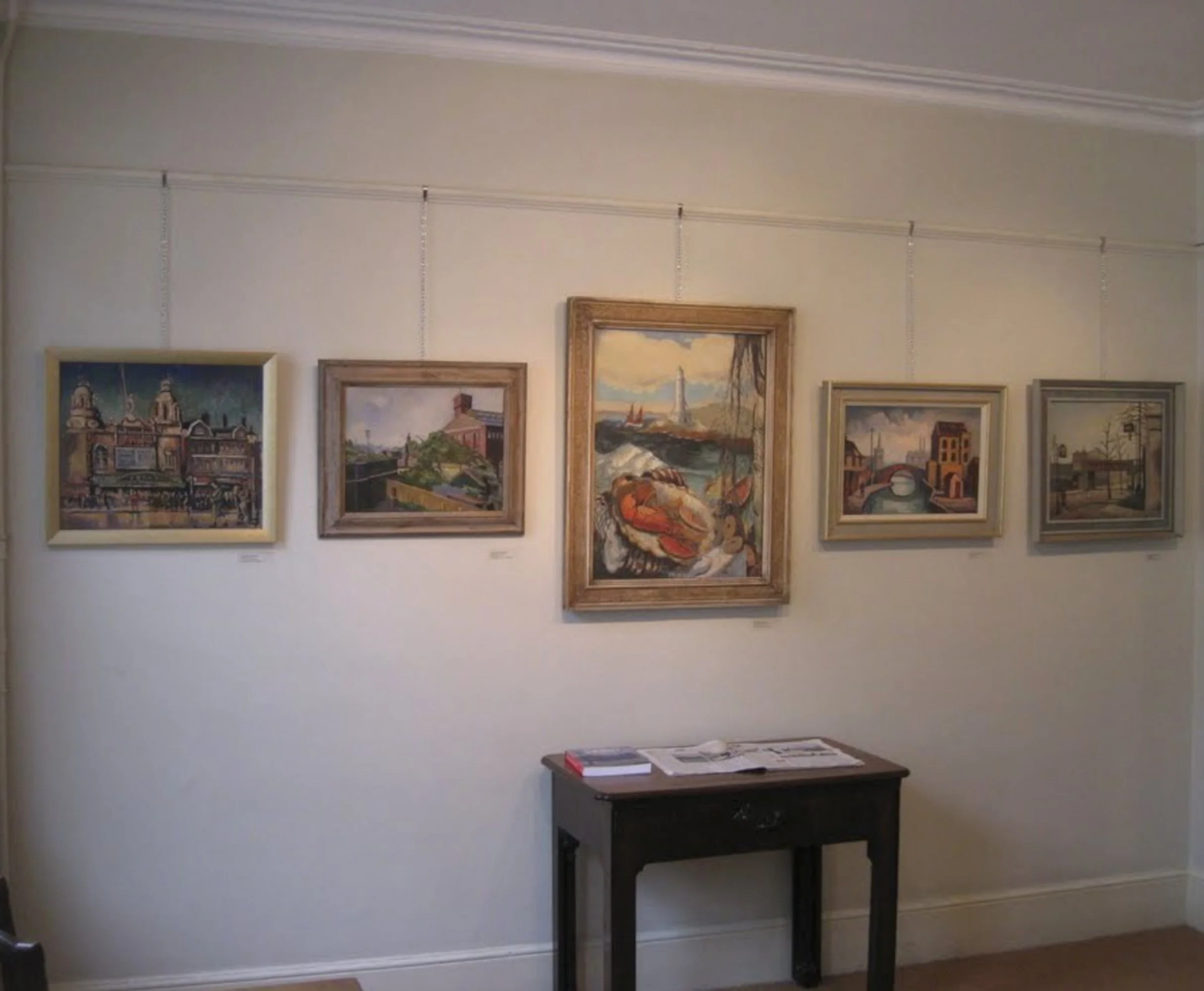 Six paintings hanging on a white wall in an art gallery, with a small wooden table underneath holding some papers and books.