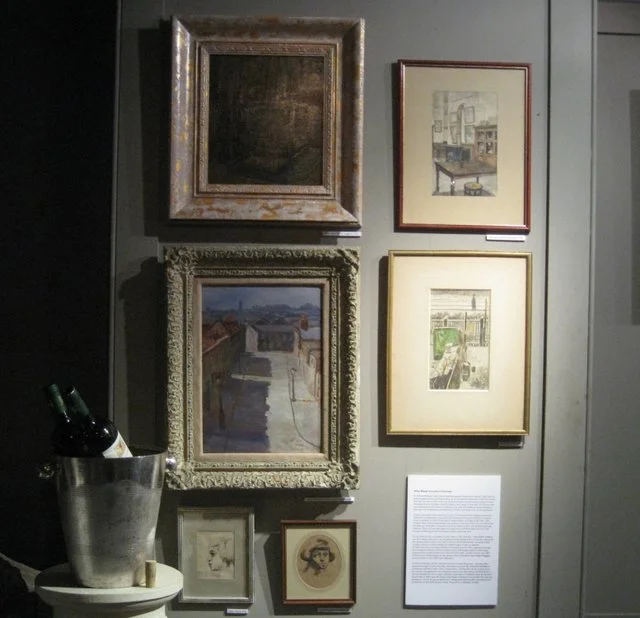 Picture of an art display wall with five framed artworks, a white informational plaque, and a metal bucket containing wine bottles.