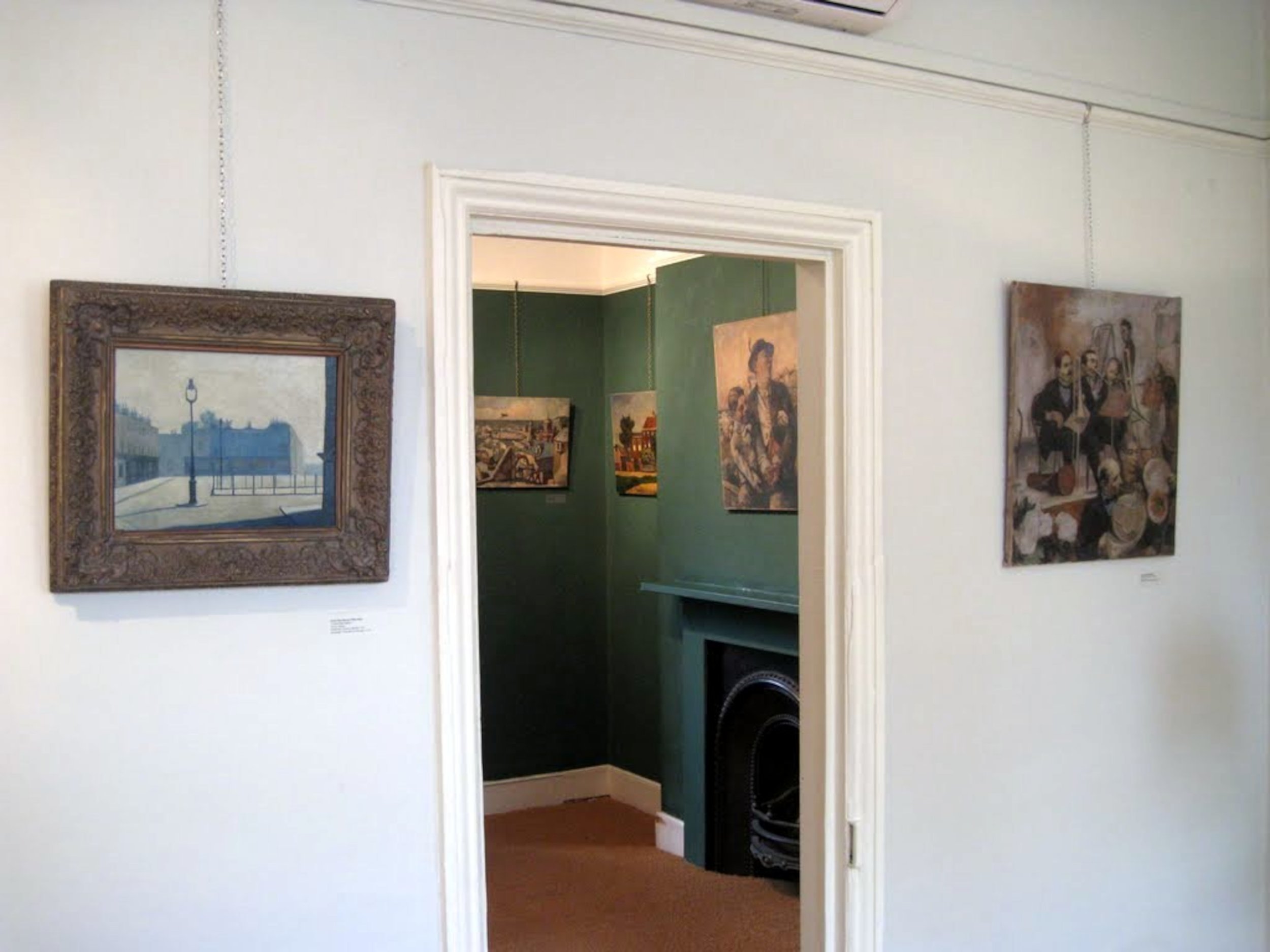 Interior view of an art gallery with paintings on white and green walls. The focus is on the white wall with three paintings, two on the left and one on the right. A doorway leads into a room with more paintings and a fireplace.