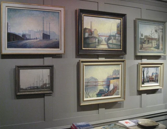 Six framed landscape paintings hanging on a gray wall, depicting industrial and urban scenes with water, bridges, and boats.