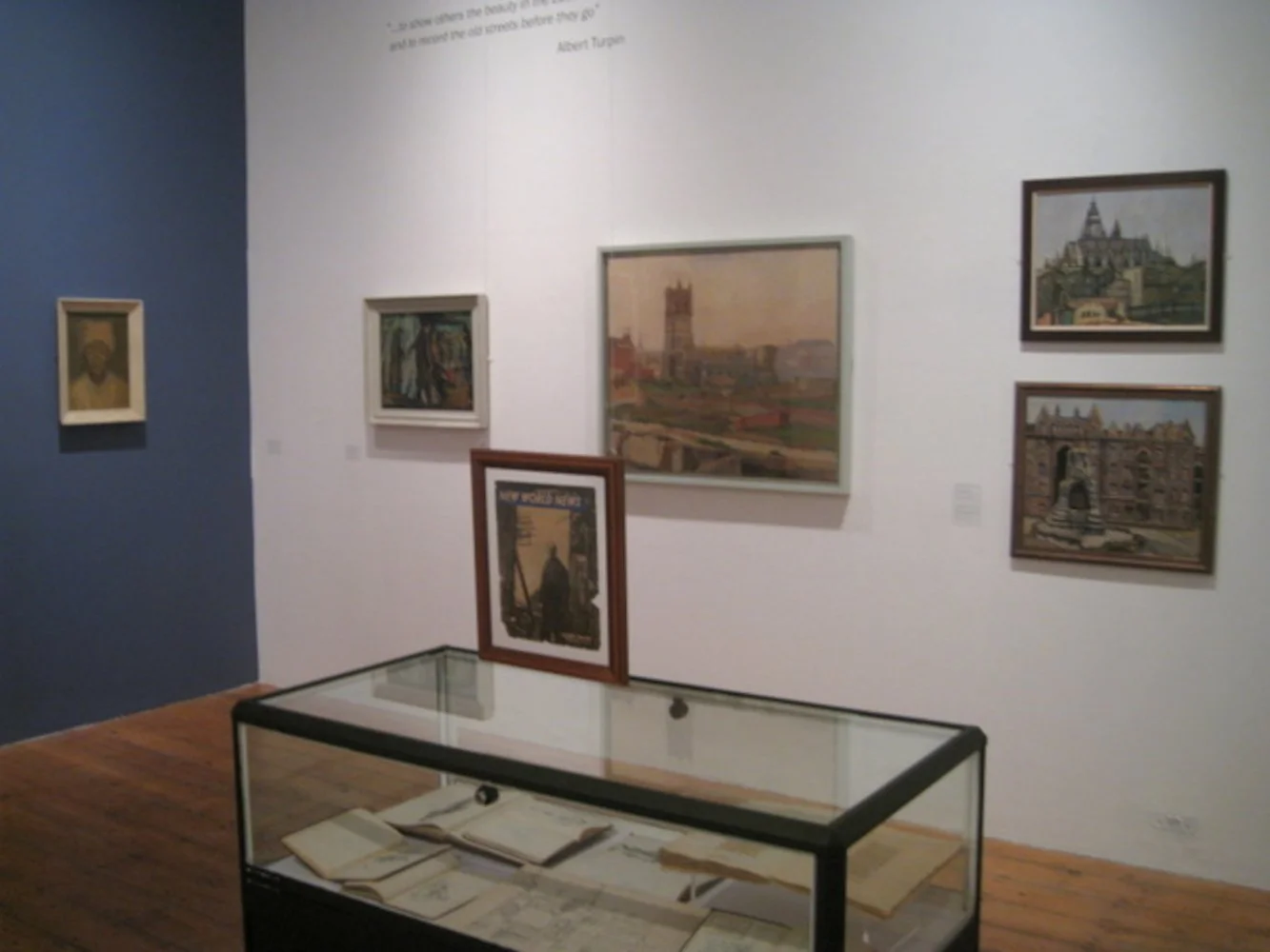 Art gallery displaying six framed paintings on white and blue walls, a glass display case with open books and a framed artwork on top.