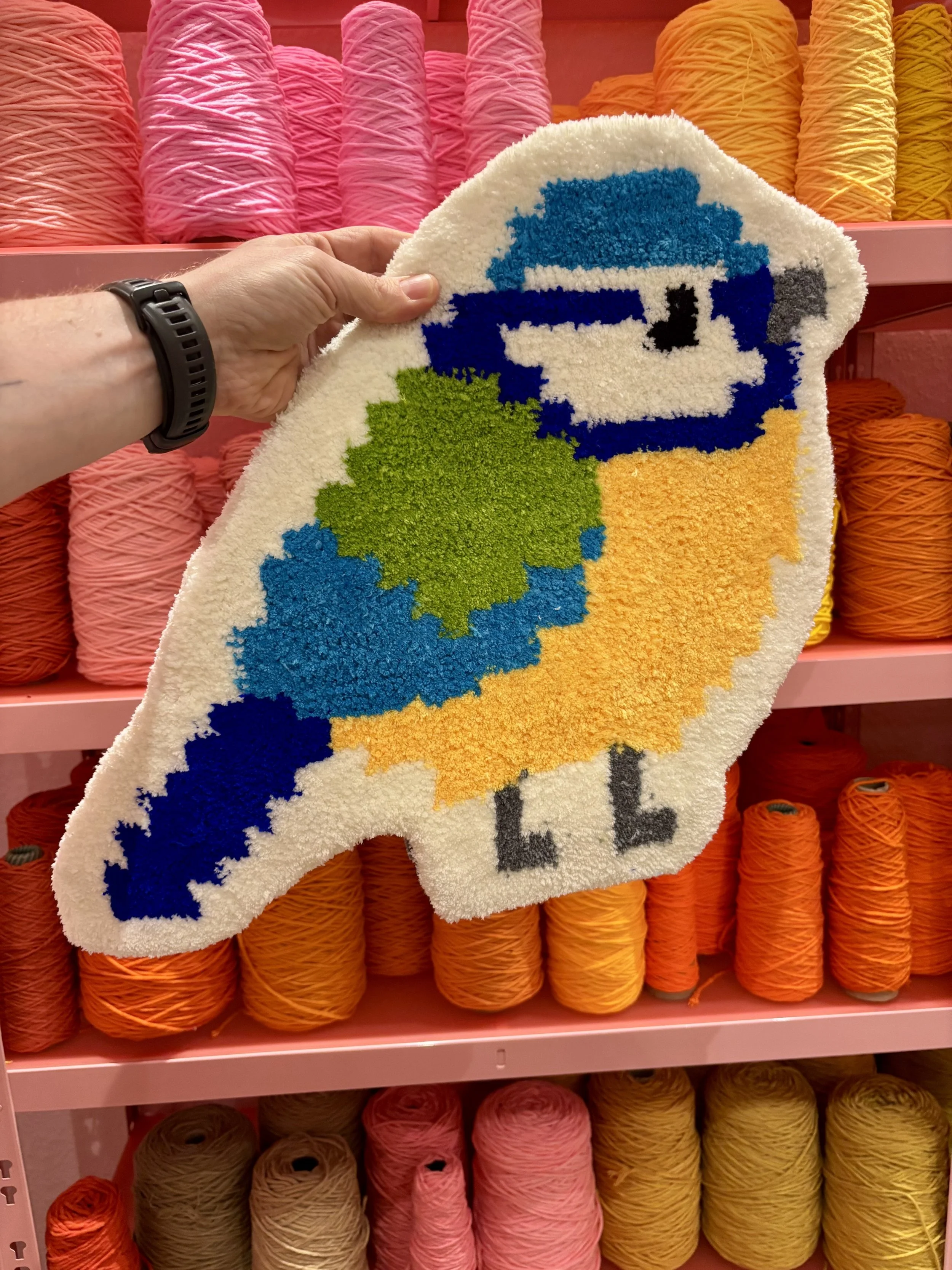 A person holding a colorful pixelated towel with a helicopter design in front of yarn spools in various shades of pink, orange, and yellow. (The OG)