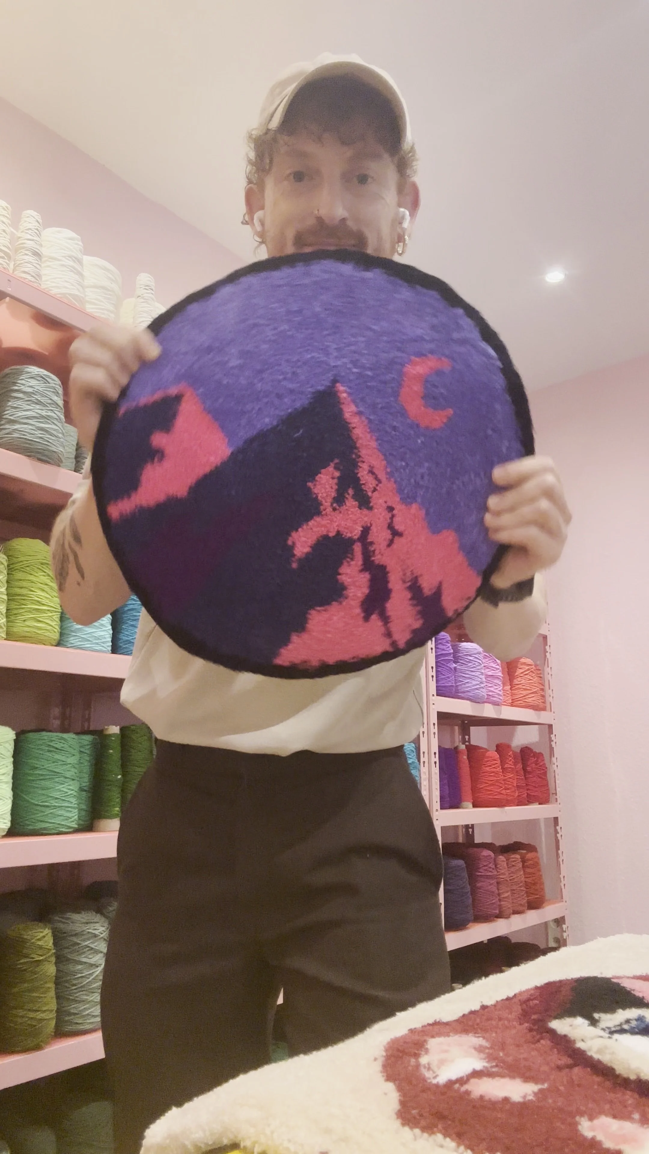 Person holding a round rug or mat with a colorful design, standing in a store with shelves of yarn in the background.