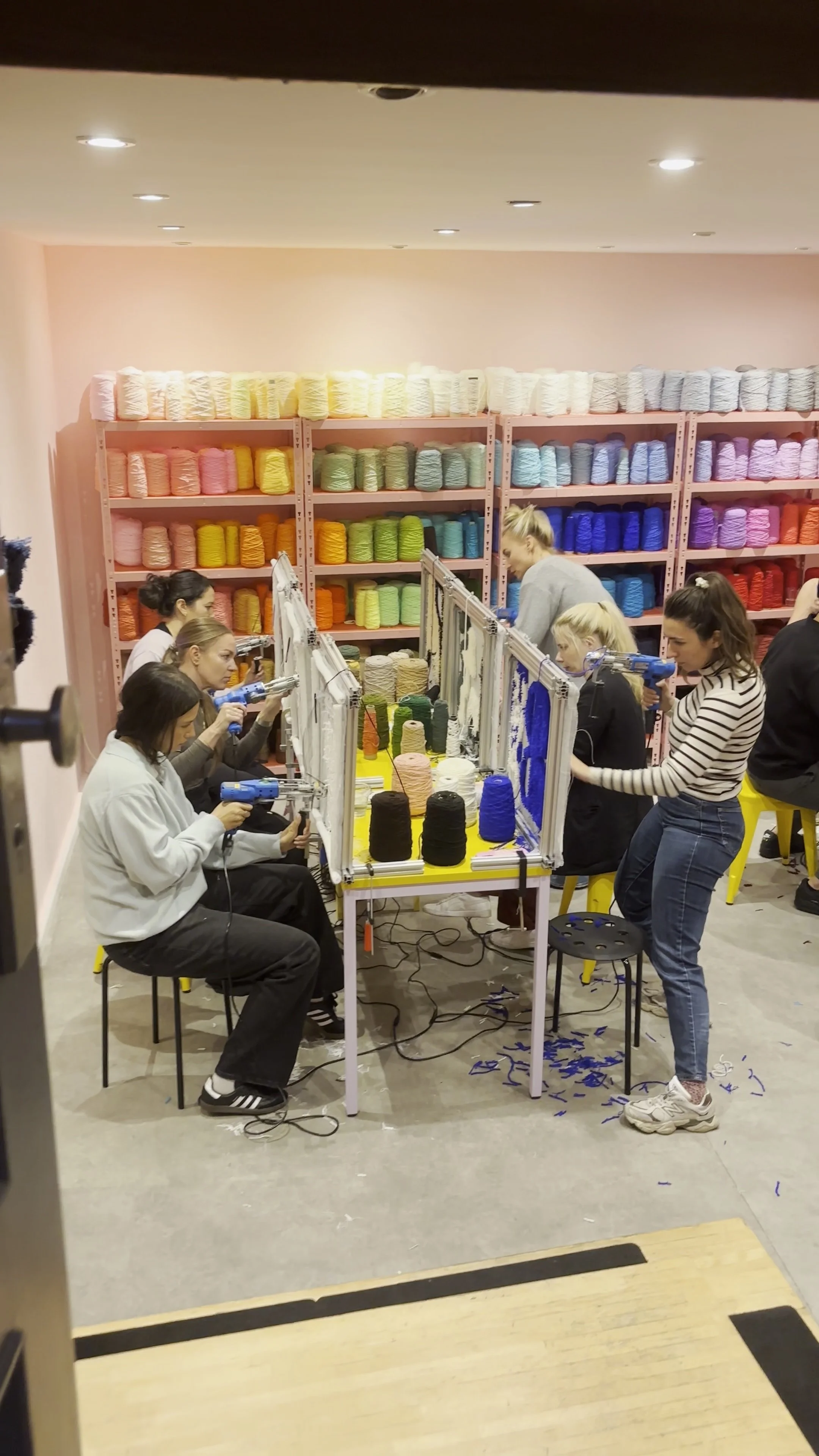 People sitting on stools around a table, engaged in a craft activity involving yarn in a colorful yarn shop with shelves filled with yarn in the background.