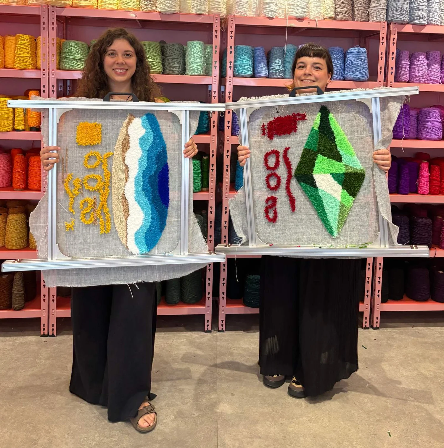 Two people hold up their tufted rugs in frames in front of the yarn wall