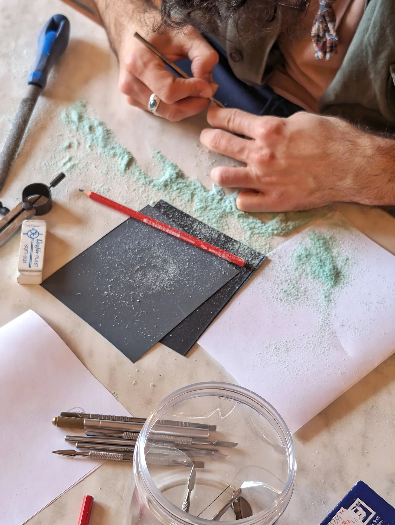 Hands carving a wax ring at a workbench, with green wax shavings and tools