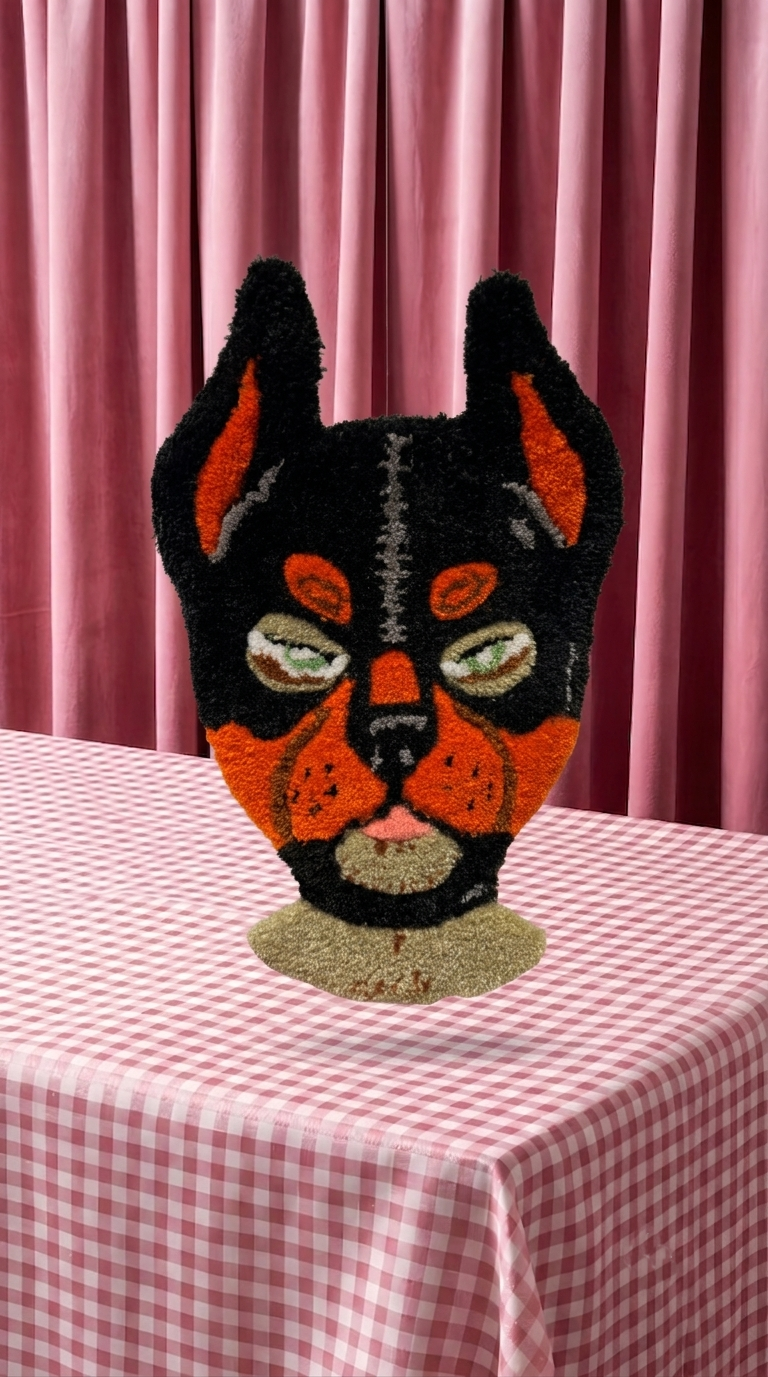 A decorative mask resembling a feline face with black, orange, and beige colors, resting on a pink-and-white checkered tablecloth with pink curtains in the background.