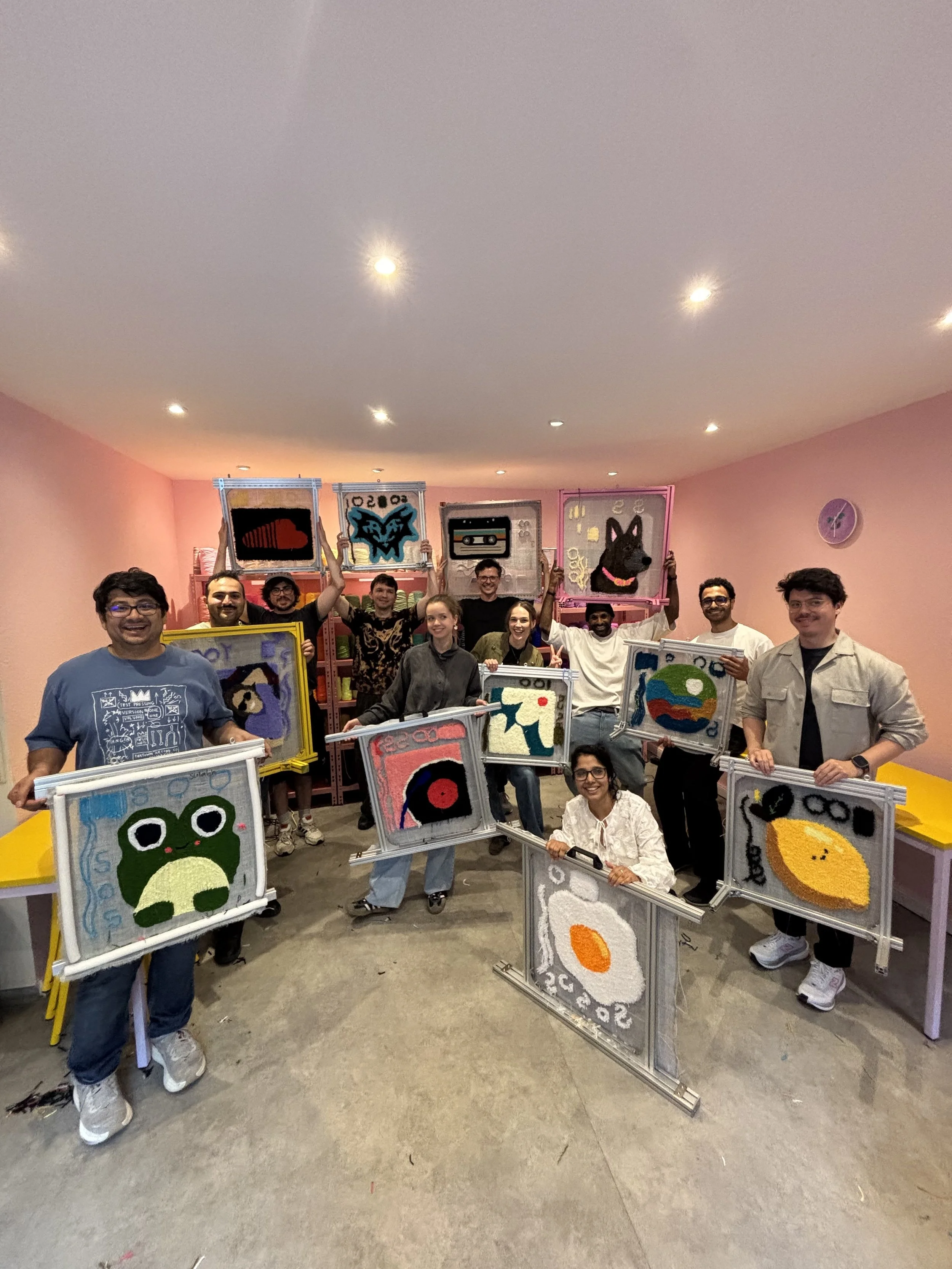 A group of people holding colorful artistic rugs with various abstract designs and bright colors, standing in a room with pink walls and a gray floor. (The OG)