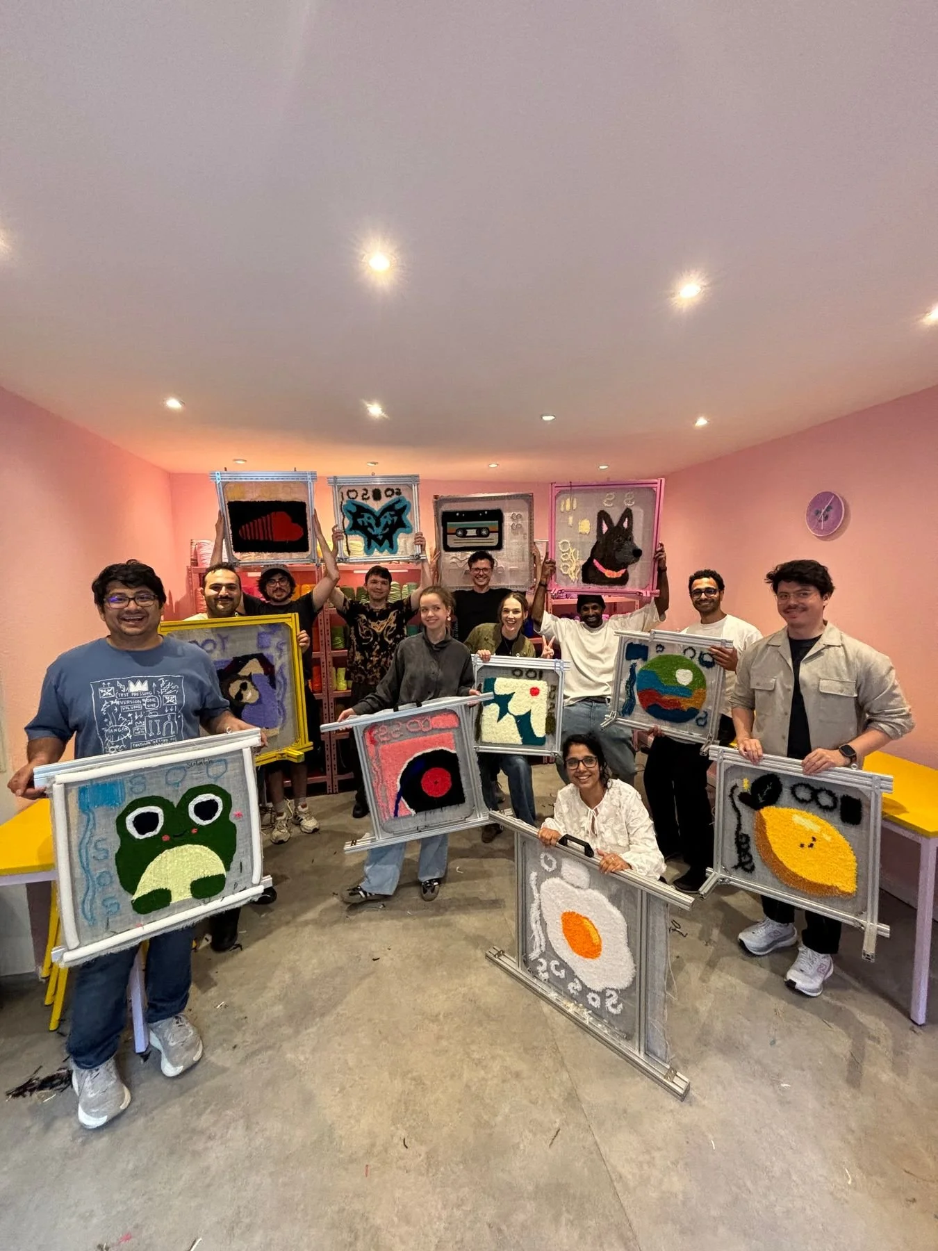 A team event where a group of people hold up their tufted artworks. They're in a beautiful pink studio space.