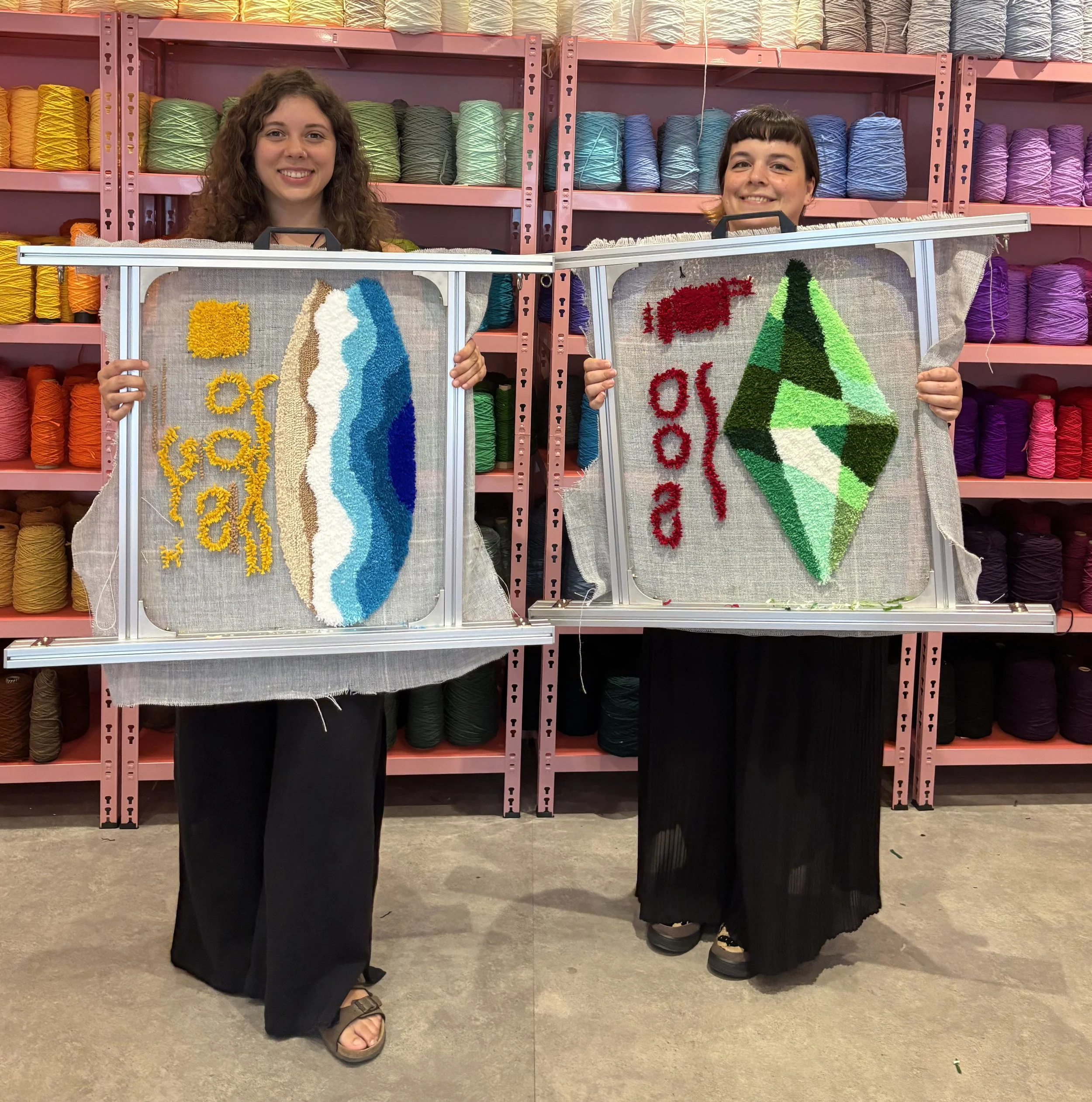 Two women standing behind a display of textile art with colorful yarn shelves in the background. (The OG)