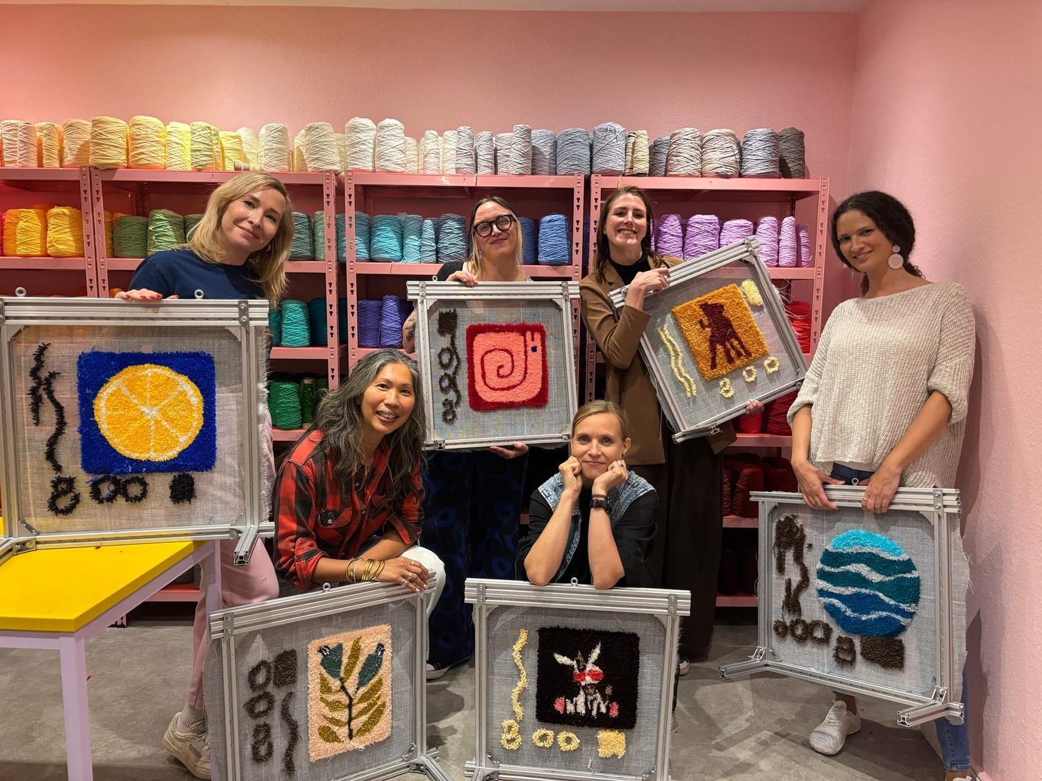 A group sits with their tufted rugs in front of the yarn wall