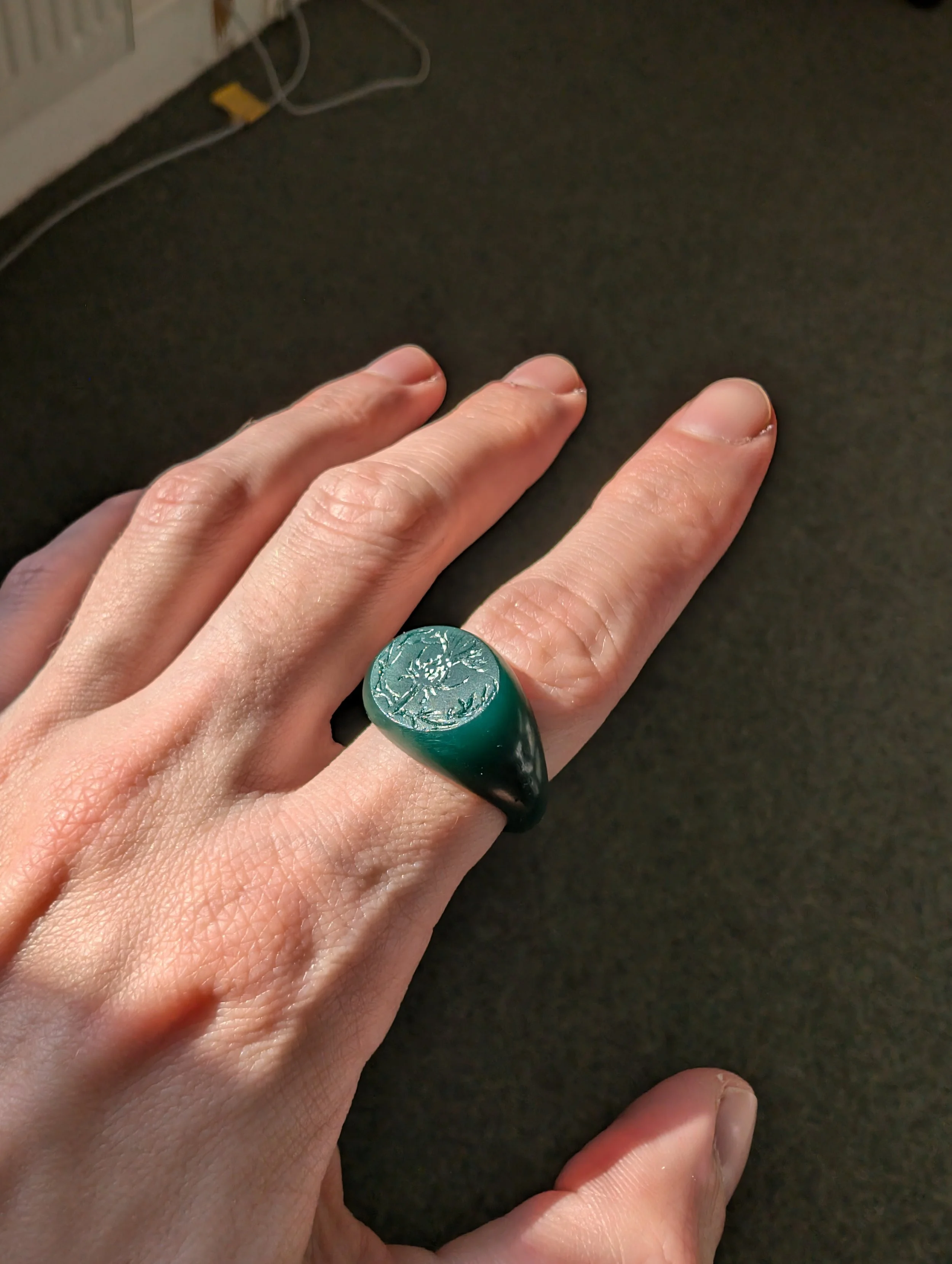 Hand wearing a green wax signet ring with an engraved design, before casting