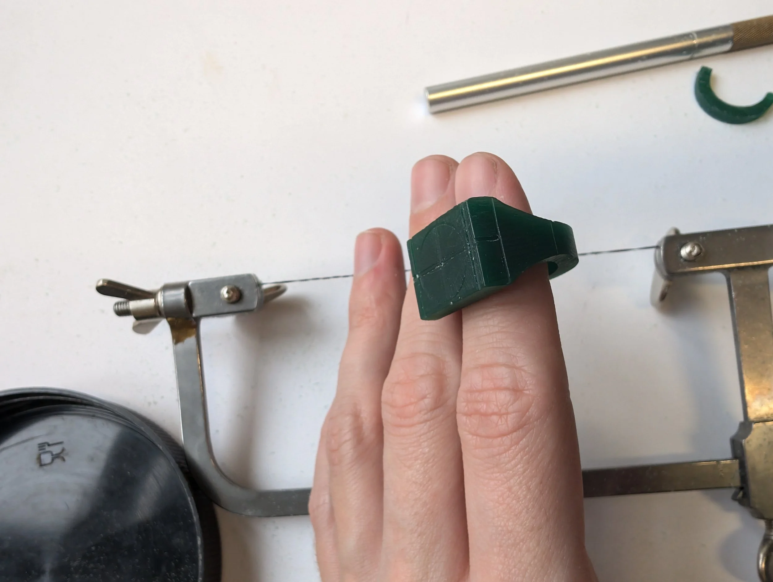Green wax ring model held against two fingers to check the fit, beside a jeweller’s saw