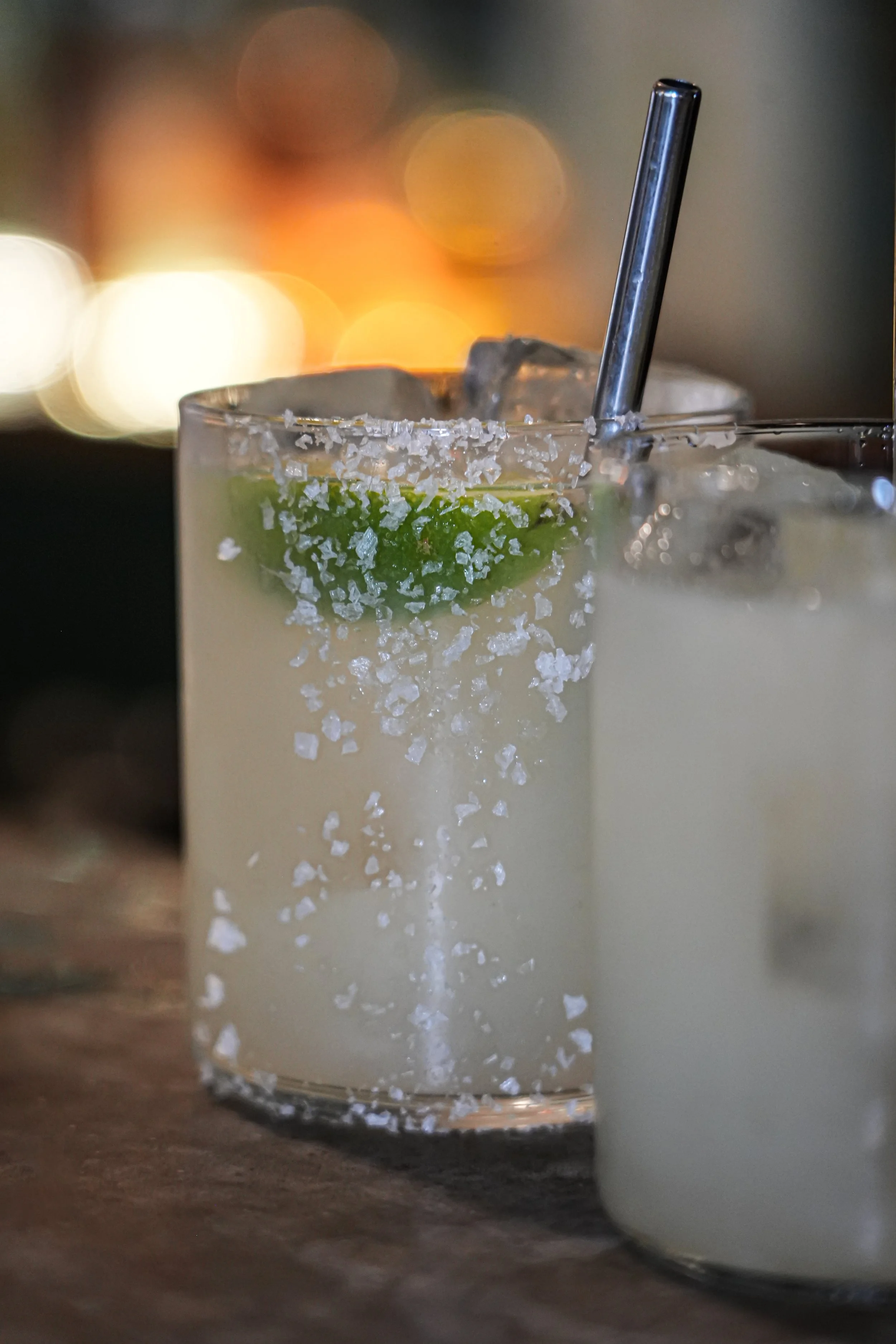 A glass of margarita with salt rim, lime wedge garnish, and a metal straw on a dark surface with a blurred colorful background.