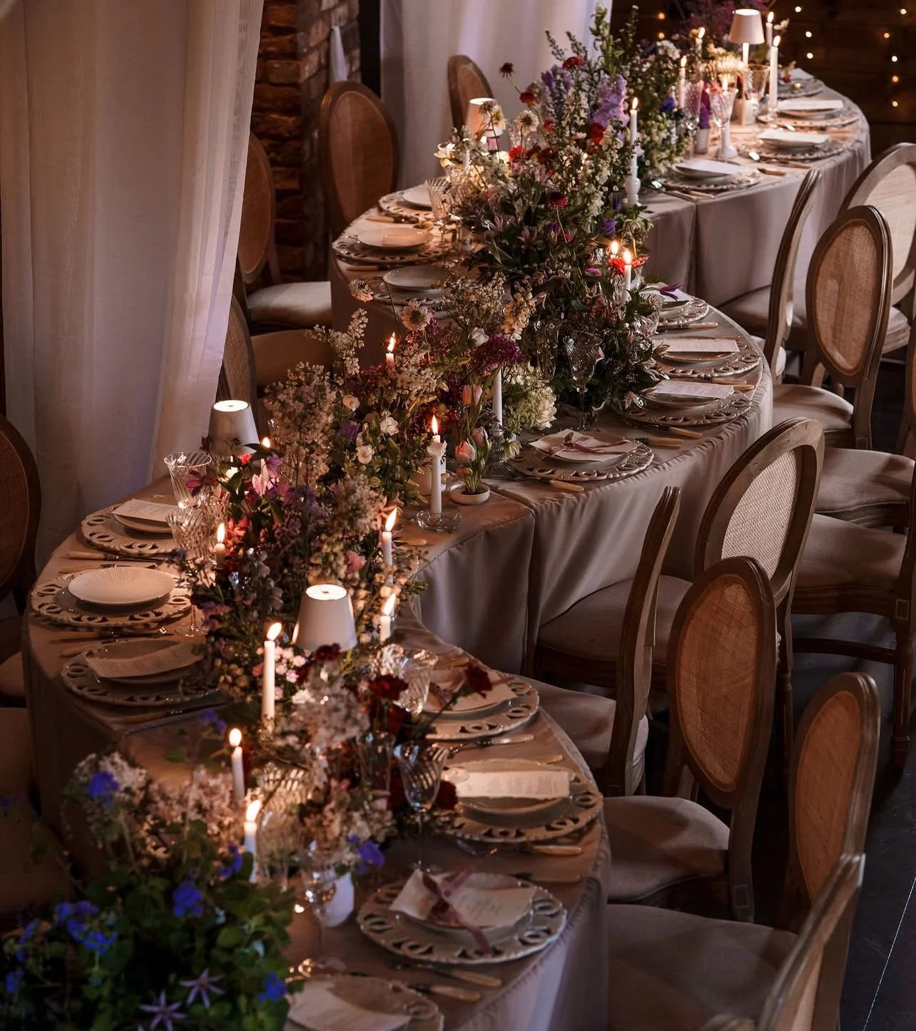 A long dining table decorated with floral arrangements, candles, and place settings for a formal event in a cozy, warmly lit room.