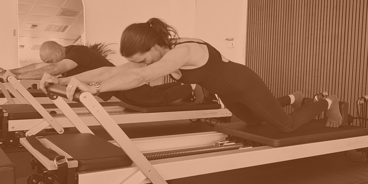 Two people are on Reformers, performing Pilates exercises with trainer assisting one woman.