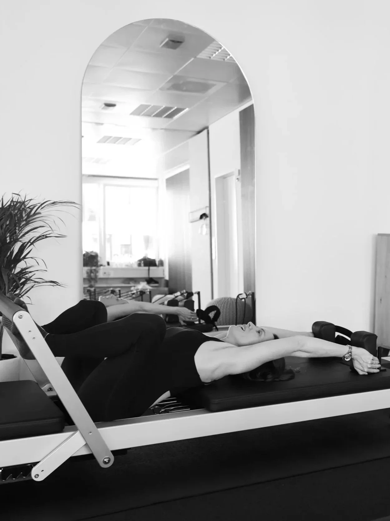 Person lying on a reformer pilates machine stretching her arms above her head during exercise, in a room with another person and fitness gear visible in the background.