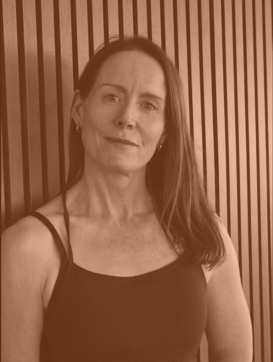 A woman with long red hair wearing a dark tank top standing in front of a wooden slat wall.