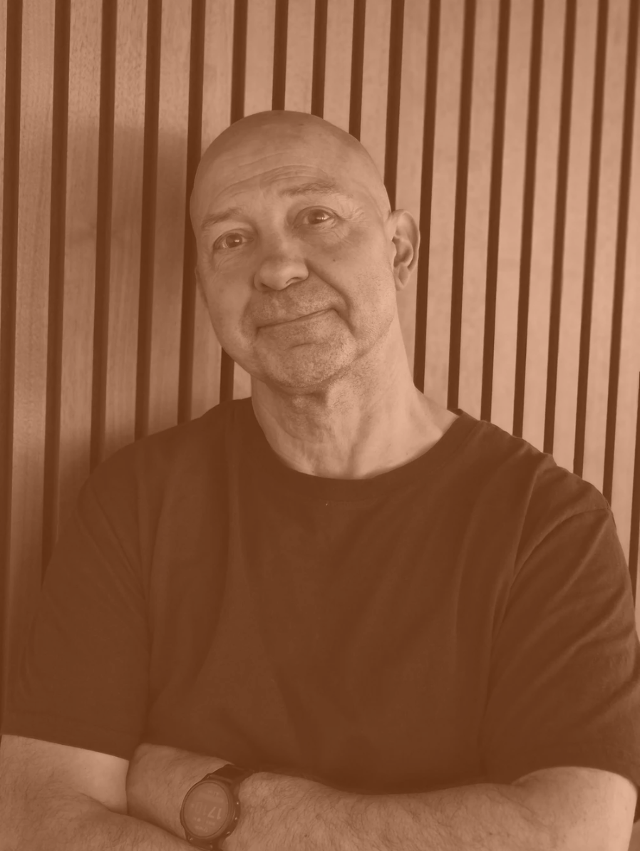 A middle-aged man with a shaved head, smiling gently, wearing a dark t-shirt, standing in front of a wooden slat wall.