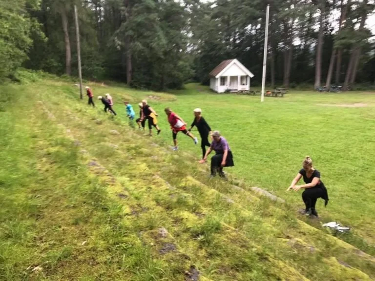 Grupper av personer, mest kvinner, som planter små planter i en skråning med gress. Det er en liten hytte i bakgrunnen, omgitt av trær.