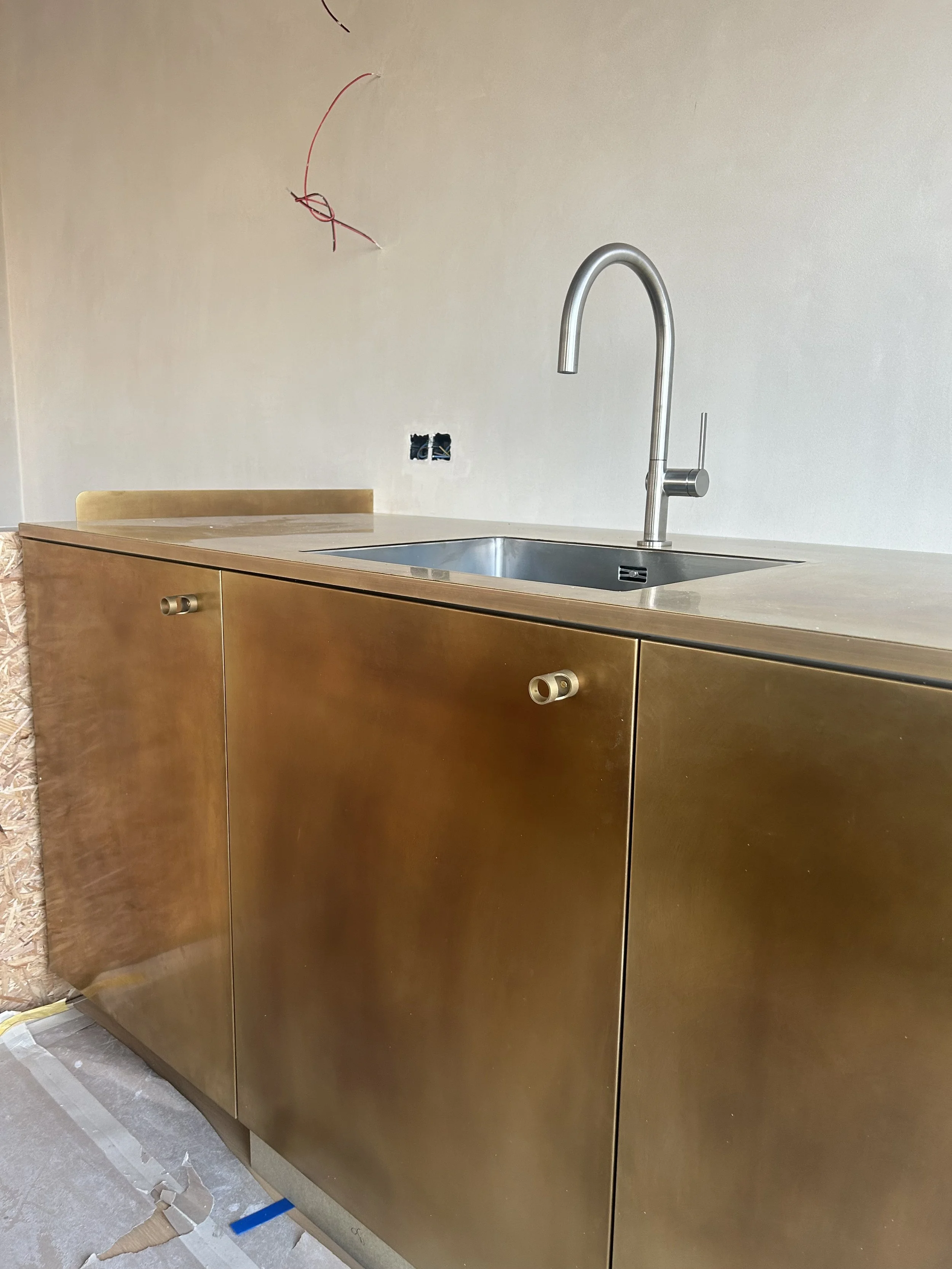 Keuken met een gouden kast, een inox gootsteen en een hoog metallisch kraan. Het plafond heeft elektrische bedrading en een open kastje zonder afwerking. Er ligt verfresten en bouwmateriaal op de vloer.