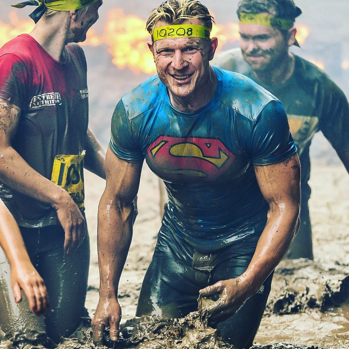 Participants covered in mud during an obstacle course race, one wearing a Superman shirt and a yellow headband with numbers.