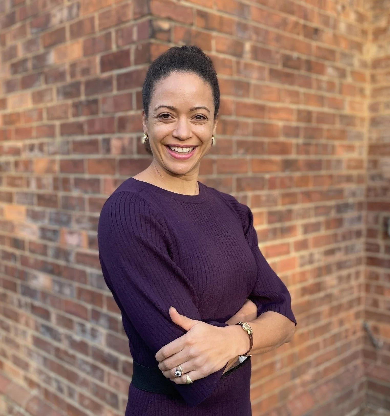 A woman with dark hair tied up, smiling and crossing her arms, standing in front of a brick wall, wearing a purple long-sleeve shirt, earrings, a bracelet, and a ring.