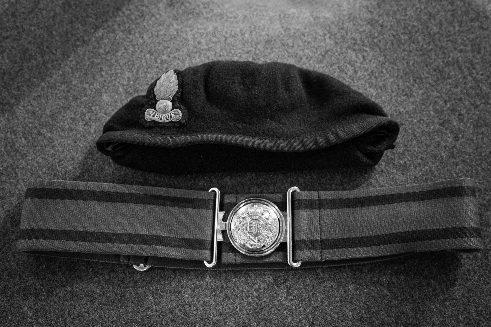 Black baseball cap with a badge that has a feather and the word 'BIGGUS' on it, resting on a striped belt with a large ornate belt buckle, laid on a grey carpet.