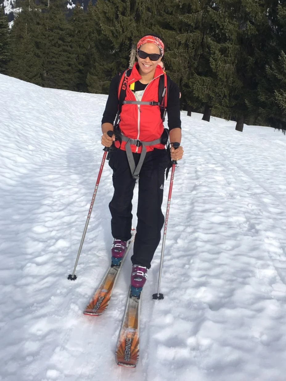 Woman in snowboarding gear, wearing sunglasses and a red jacket, standing with snow skis in a snowy forested area, smiling.