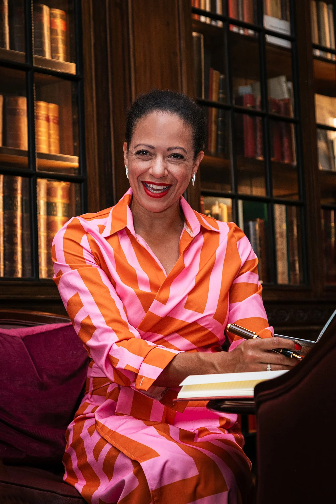 A woman in a bright orange and pink striped dress smiling while playing the piano in a room with dark wooden bookshelves filled with books.