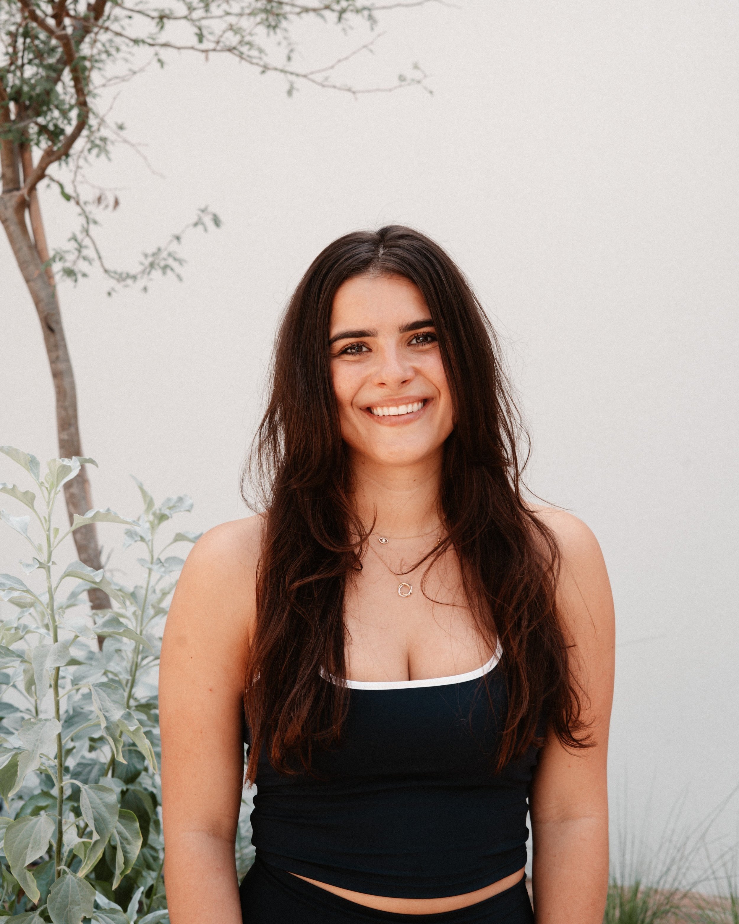 A young woman with long dark hair and light skin, smiling, standing outdoors against a plain light wall, wearing a black sleeveless top and layered necklaces.