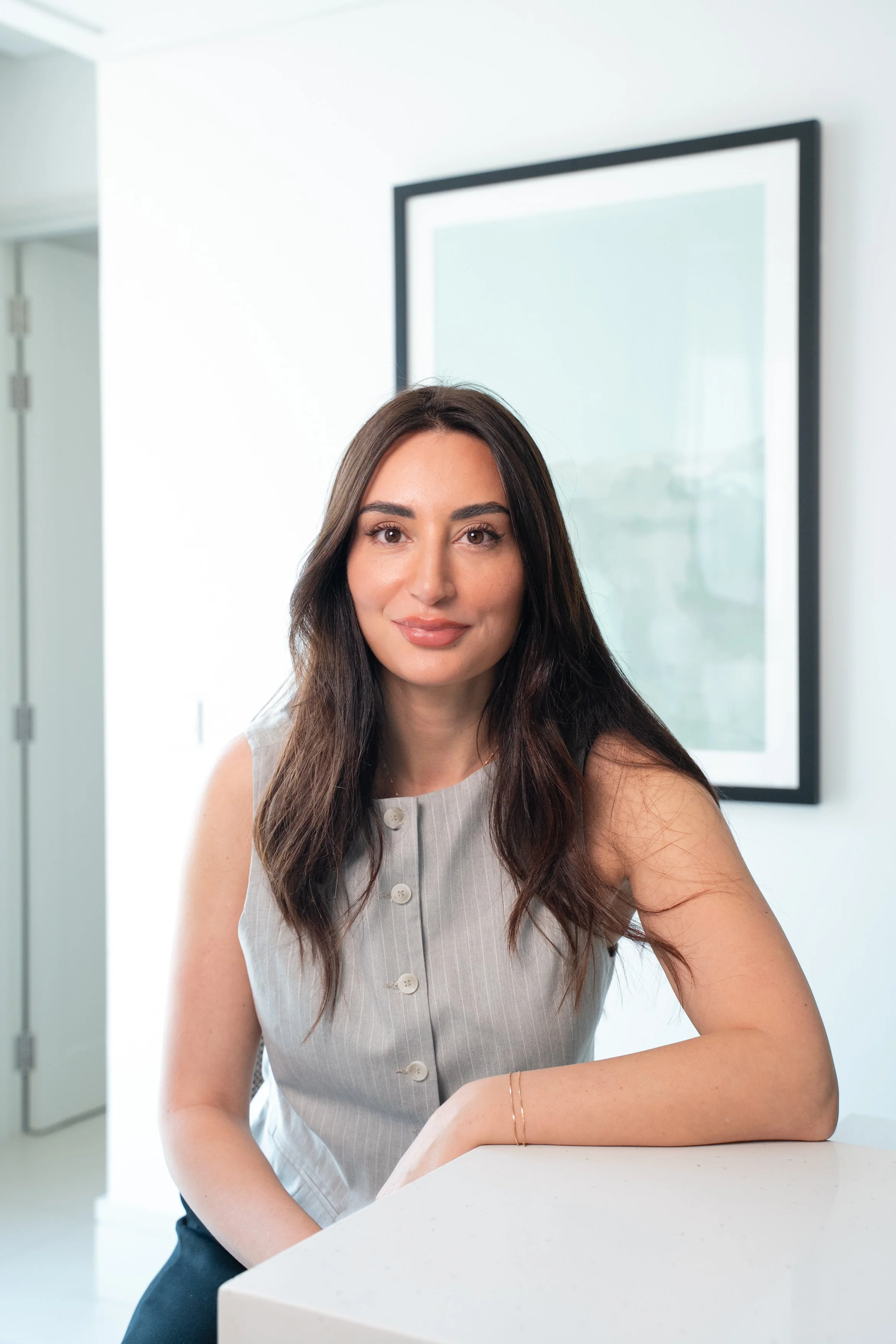 A woman with long dark hair and a sleeveless gray top sitting at a white counter in a modern, brightly lit room with minimal decor.
