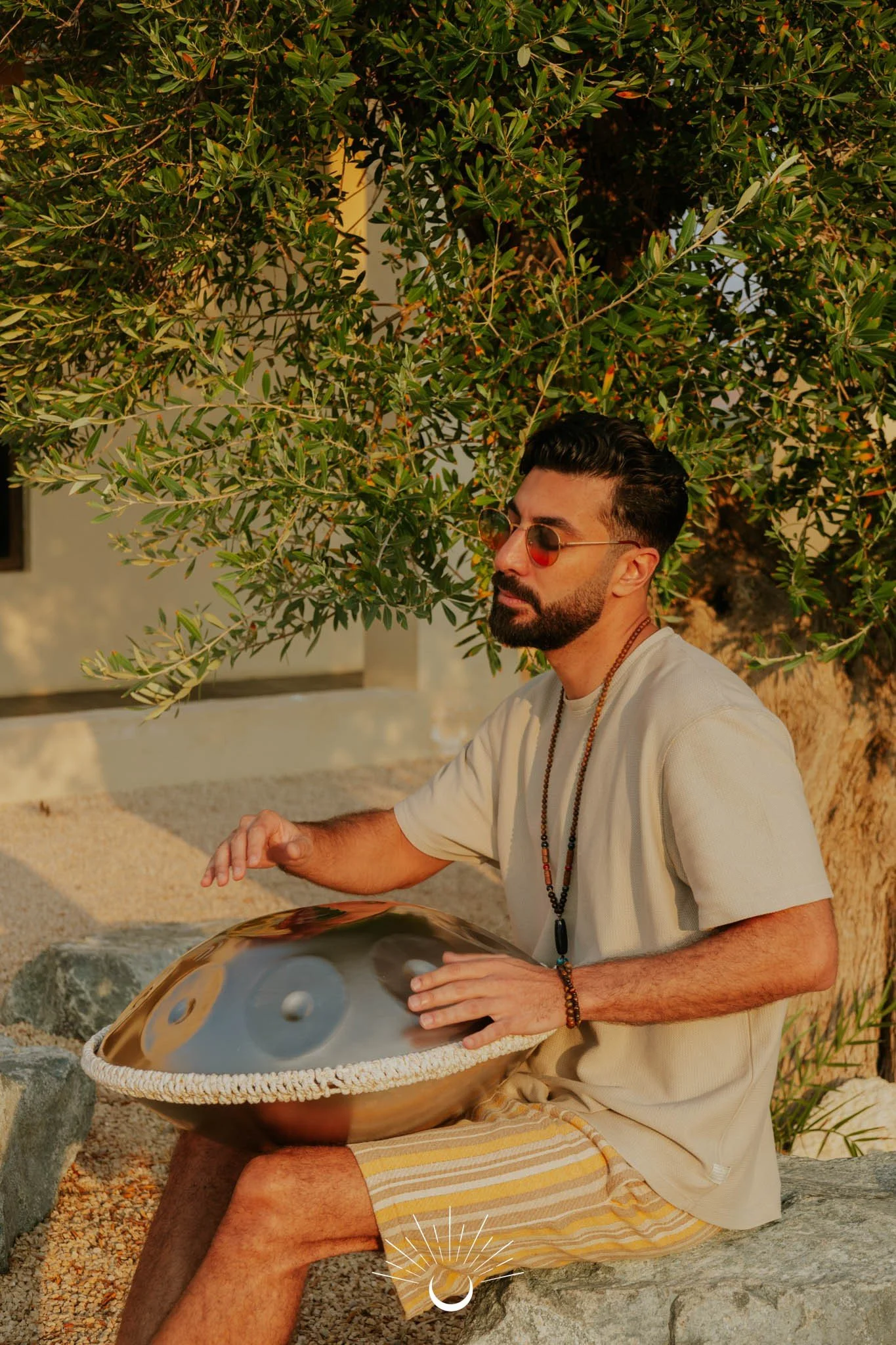 A man with dark hair and beard, wearing round sunglasses, a cream shirt, and striped shorts, sitting on a rock outdoors under a tree, playing a handpan musical instrument.
