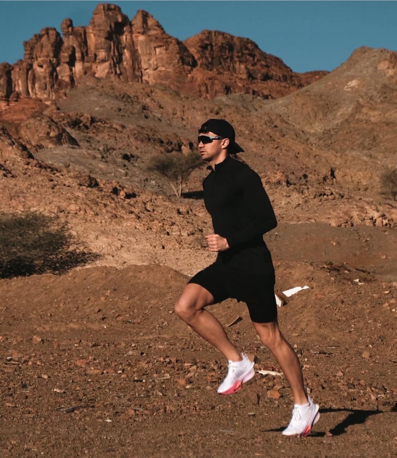 A man running in a desert landscape with rocky mountains in the background, wearing black athletic clothing, white running shoes, sunglasses, and a backward cap.