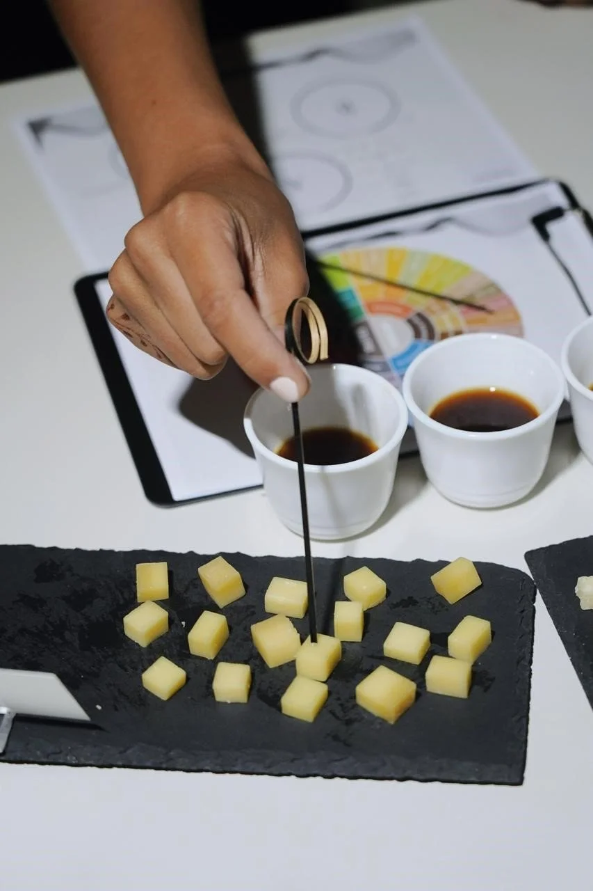 Person tasting cheese with a toothpick, grilled cheese on black slate, three cups of dark liquid, and a color wheel chart in background.
