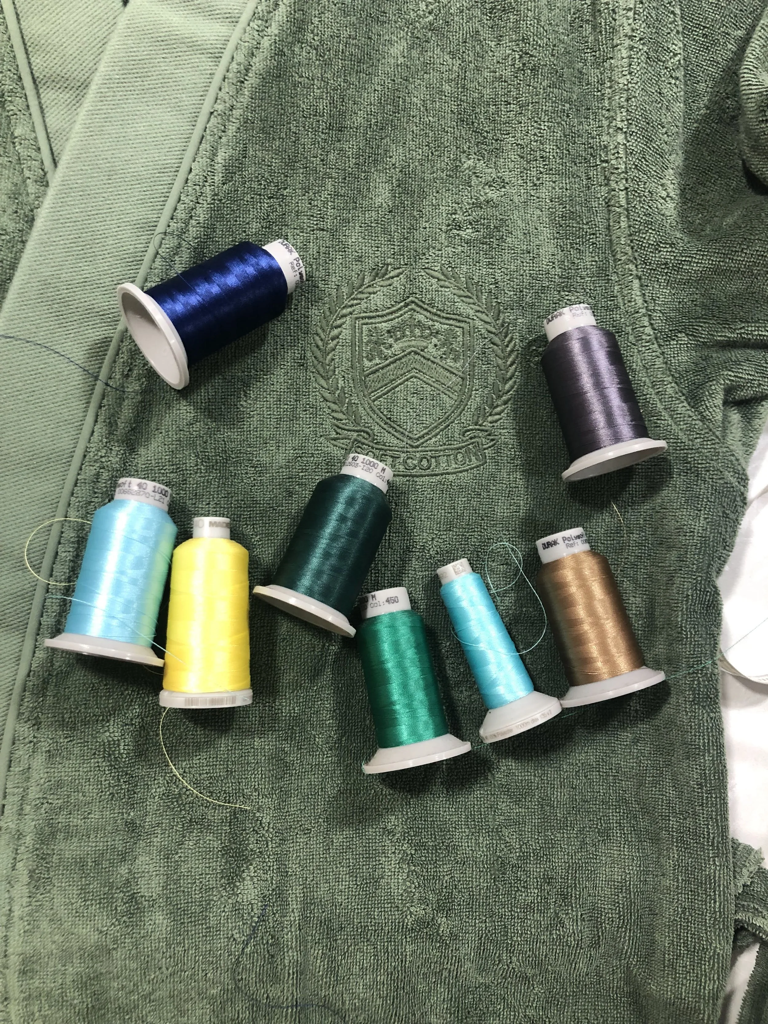 Multiple spools of sewing thread in various colors, including blue, yellow, green, turquoise, purple, and gold, placed on a green towel with a logo embroidered on it.