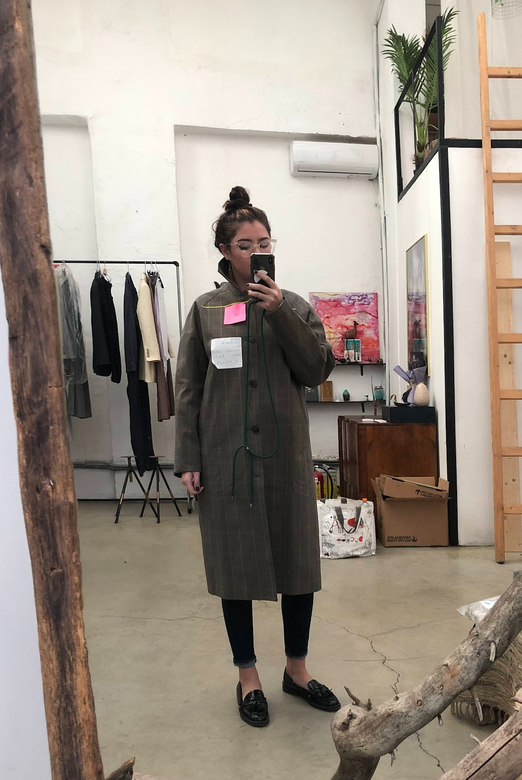 A woman taking a mirror selfie in a store or studio with clothing racks, artwork, and decorative items in the background. She is wearing glasses, a long plaid coat, black leggings, and black loafers.