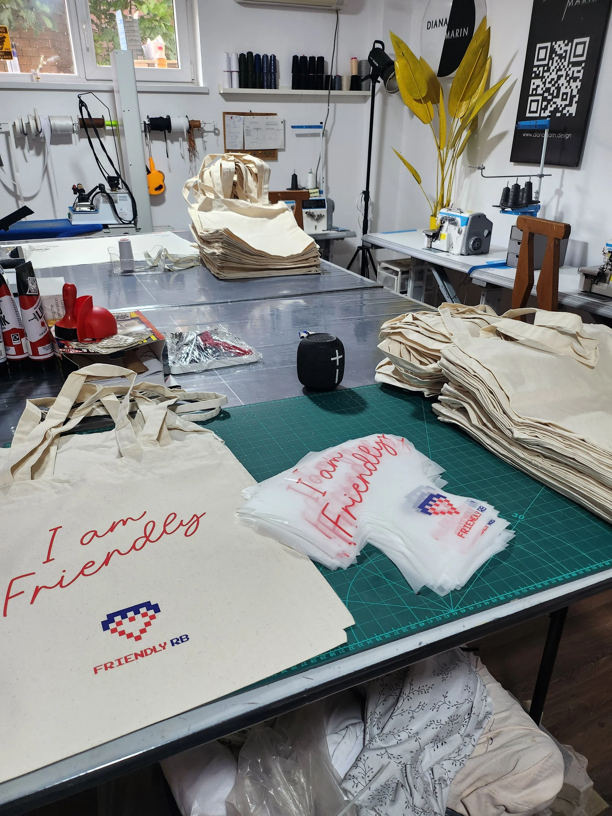 A workspace with fabric tote bags on a cutting mat, some completed with red handwritten text"I am Friendly" and a pixelated heart. The background features shelves with sewing supplies, a window, and a tall yellow plant.