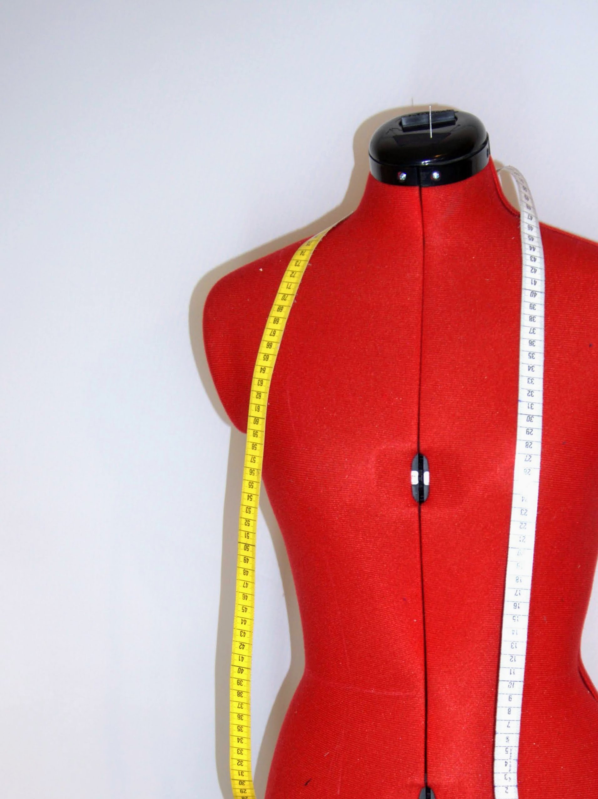 Red dress form with a black neck cap, measuring tapes draped over the shoulder and down the front, set against a plain white background.