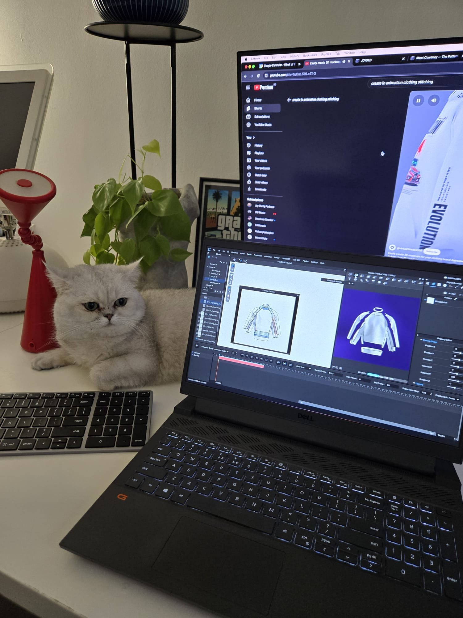 A workspace with a laptop open to animation software, a second monitor displaying YouTube, a white cat lying on the desk, a desktop keyboard, a houseplant, and a red device with a round top.